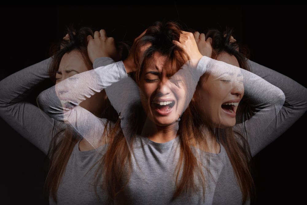 A composite photograph of a young woman experiencing extreme distress, with multiple translucent layers of her face screaming and pulling at her hair, symbolizing a panic attack or overwhelming anxiety against a dark background.