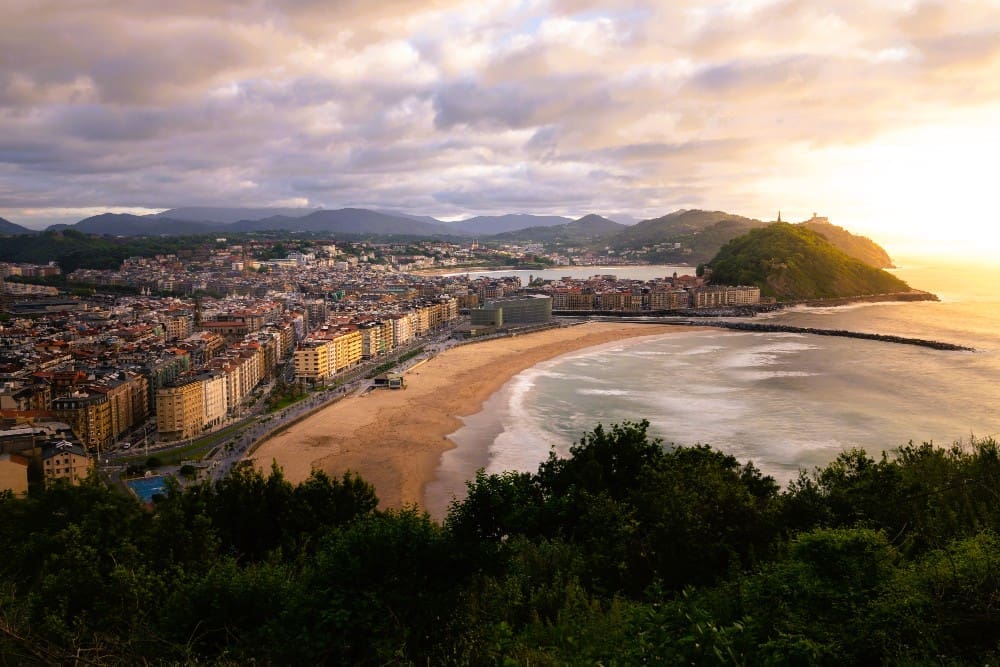 A high-angle panoramic view of the crescent-shaped La Concha Bay in San Sebastian, Spain, with a city lining the shore, a prominent hill with a statue on the right, and distant mountains under a dramatic sunset sky.