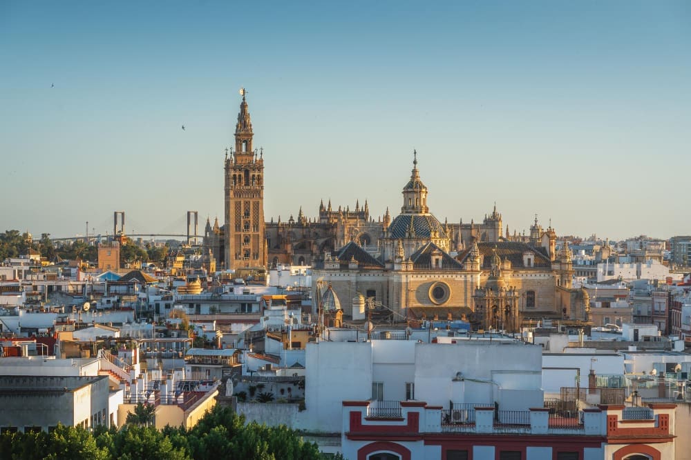 An expansive panoramic view of Seville, Spain, showcasing its iconic cathedral and Giralda bell tower towering over a dense cluster of traditional white and terracotta buildings under a clear blue sky.