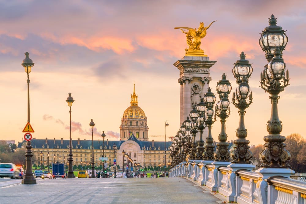 A view of Pont Alexandre III bridge with ornate lampposts leading towards Les Invalides dome in Paris at sunset.