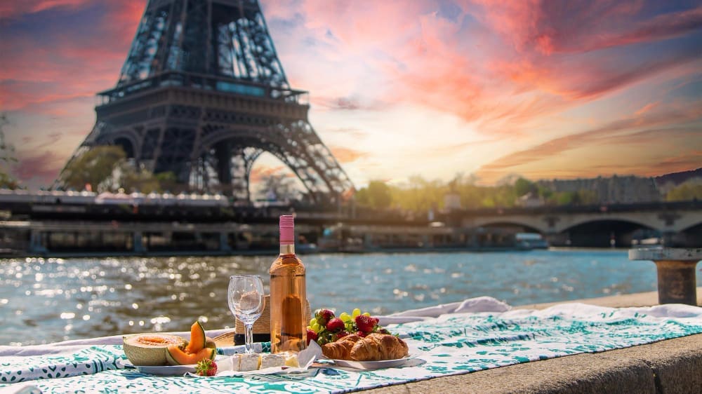 A close-up of a picnic spread with a bottle of rosé wine, wine glasses, a croissant, strawberries, and melon, set on a blanket by the Seine River, with the Eiffel Tower in the blurred background at sunset.