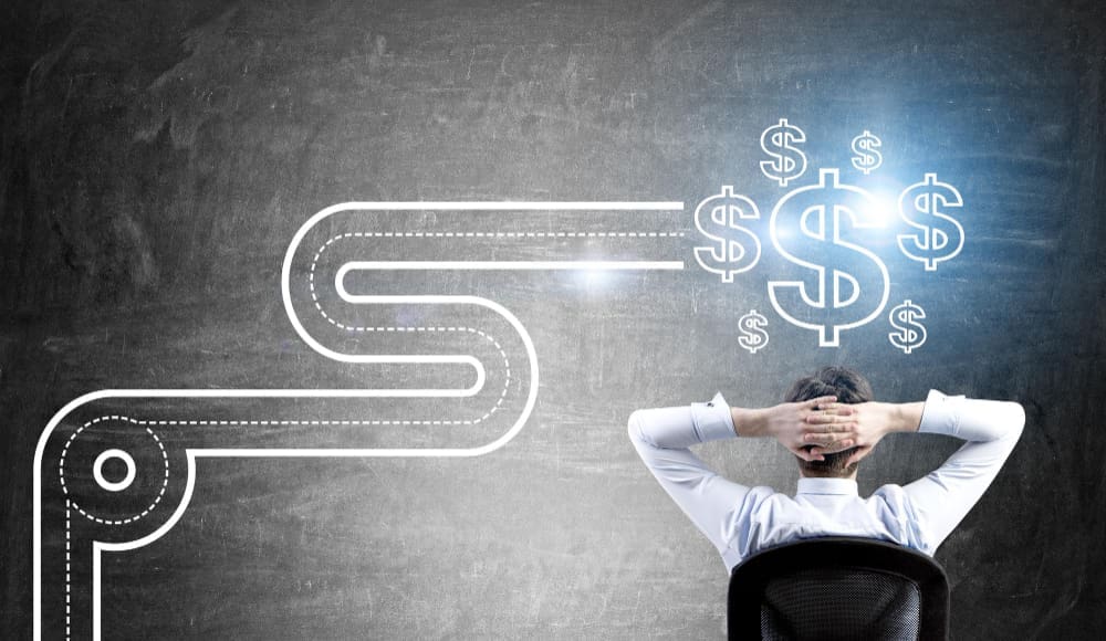 A man in a white shirt, seen from behind, sits in a chair with his hands behind his head, contemplating a winding road drawn on a grey wall that leads to glowing dollar signs.