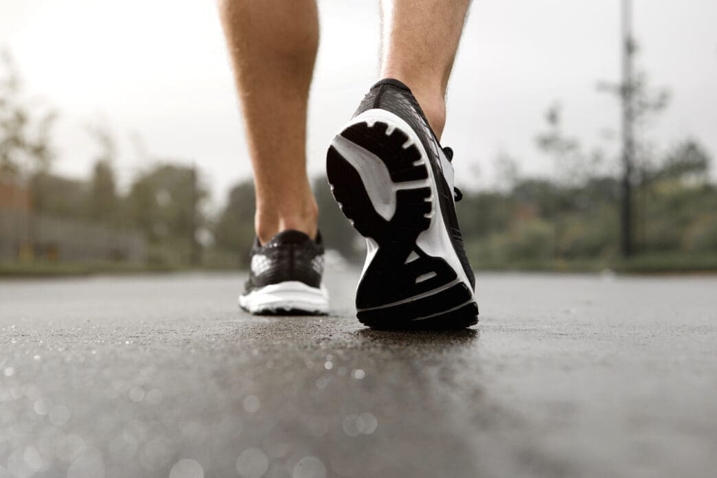Close-up of a person's feet in black and white running shoes walking on a wet paved surface, seen from behind.