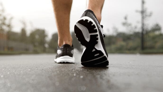 Close-up of a person's feet in black and white running shoes walking on a wet paved surface, seen from behind.