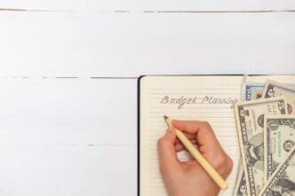 A person's hand writes "Budget Planning" in a notebook, with U.S. dollar banknotes tucked inside, all placed on a white wooden surface.