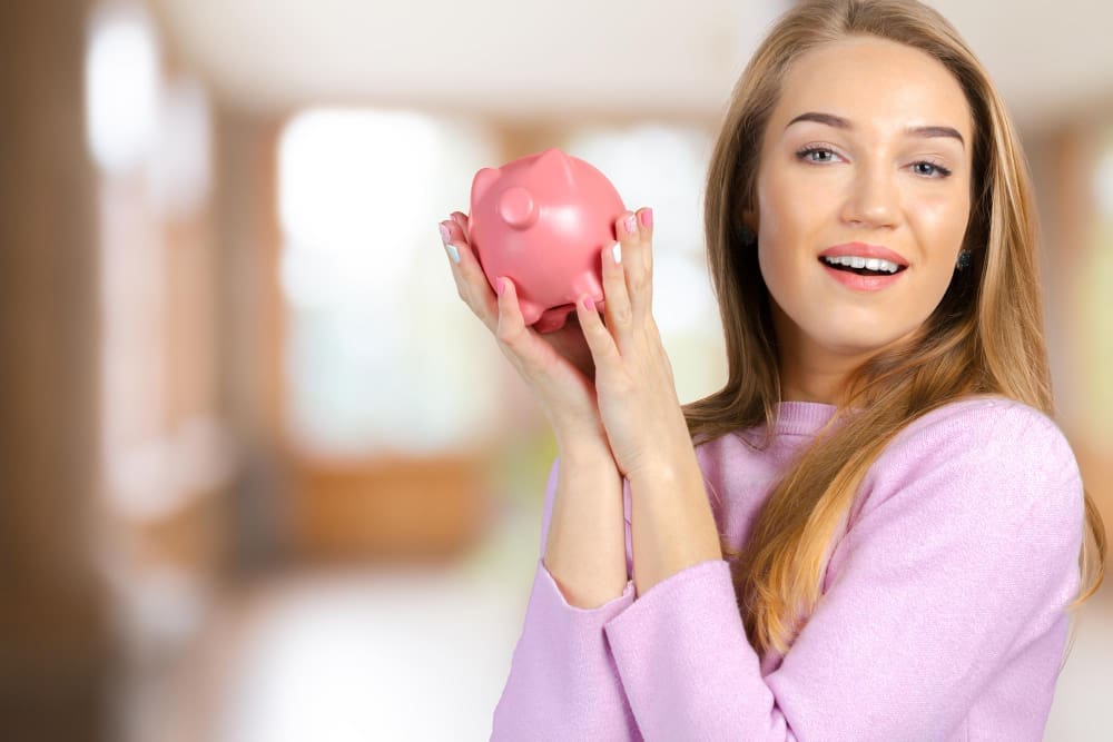 A happy young woman with long blonde hair, holding up a pink piggy bank with both hands, smiling widely at the camera, symbolizing personal finance and saving money.