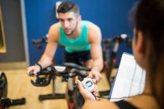 A personal trainer's hand holds a stopwatch and clipboard, timing a male client who is actively exercising on a stationary bike in a gym setting.