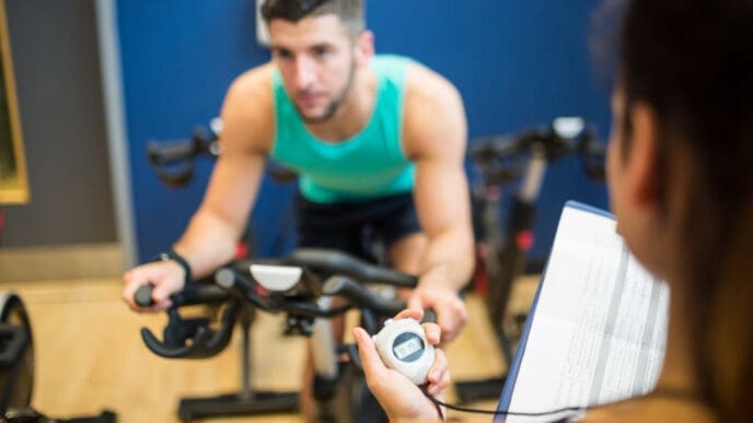 A personal trainer's hand holds a stopwatch and clipboard, timing a male client who is actively exercising on a stationary bike in a gym setting.