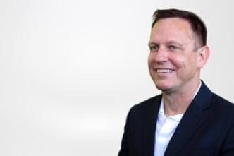 A professional headshot of a smiling Peter Thiel with short dark hair, wearing a dark suit jacket and a white shirt, looking off to the right against a plain white background.