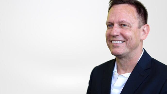 A professional headshot of a smiling Peter Thiel with short dark hair, wearing a dark suit jacket and a white shirt, looking off to the right against a plain white background.
