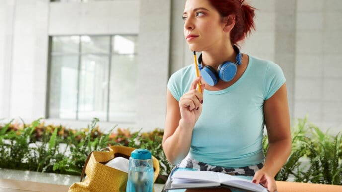A young woman with reddish-orange hair, wearing a light blue t-shirt and headphones around her neck, sits outdoors, looking thoughtfully up. She holds a pencil to her chin and an open notebook, with a water bottle and yoga mat nearby.