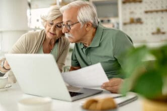 A senior couple with white hair, wearing glasses, intently looking at a laptop screen and reviewing papers together at a table, likely managing their finances.