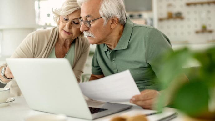 A senior couple with white hair, wearing glasses, intently looking at a laptop screen and reviewing papers together at a table, likely managing their finances.
