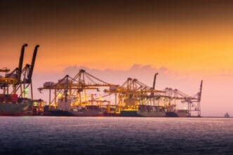 The silhouettes of numerous large gantry cranes and container ships are visible at a busy port at dusk, with the sky a beautiful gradient of orange, yellow, and purple.