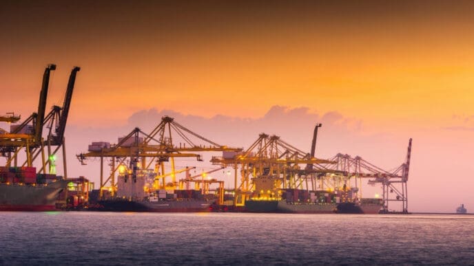 The silhouettes of numerous large gantry cranes and container ships are visible at a busy port at dusk, with the sky a beautiful gradient of orange, yellow, and purple.