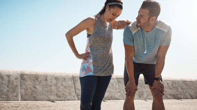 Two exhausted runners taking a break, one leaning on the other, suggesting fatigue or a potential injury.