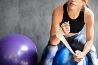 A woman in activewear wrapping her hand with athletic tape while sitting on a yoga ball in a gym.