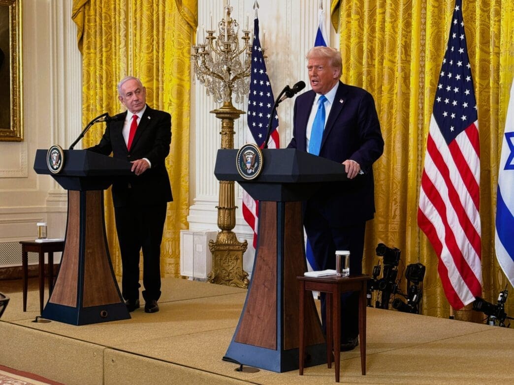 President Donald Trump and Prime Minister Benjamin Netanyahu stand at podiums with presidential and governmental seals, speaking to a press conference, flanked by American and Israeli flags in a room with golden curtains.