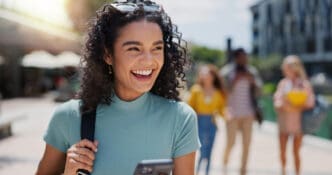 A beautiful, smiling Latina student with curly dark hair, wearing a teal top and sunglasses on her head, looks to her left while holding a smartphone. She is walking on a sunny university campus, with other students blurred in the background.