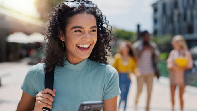 A beautiful, smiling Latina student with curly dark hair, wearing a teal top and sunglasses on her head, looks to her left while holding a smartphone. She is walking on a sunny university campus, with other students blurred in the background.