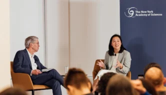 Priscilla Chan, wearing a light-colored suit, sits on a chair gesturing as she speaks, facing Dr. Richard Lifton, who is seated and looking at her, during a "fireside chat" event with the New York Academy of Sciences logo in the background.