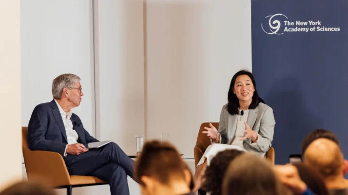 Priscilla Chan, wearing a light-colored suit, sits on a chair gesturing as she speaks, facing Dr. Richard Lifton, who is seated and looking at her, during a "fireside chat" event with the New York Academy of Sciences logo in the background.