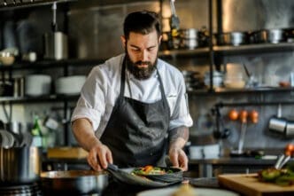 A male chef with a beard in a white chef coat and black apron, meticulously plating a dish in a professional kitchen.