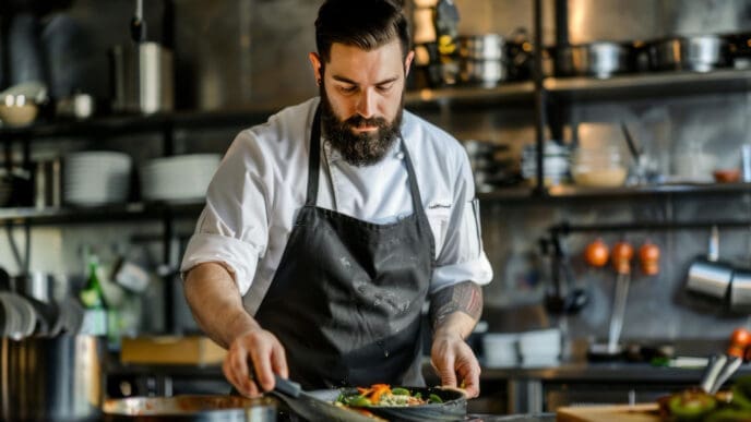 A male chef with a beard in a white chef coat and black apron, meticulously plating a dish in a professional kitchen.