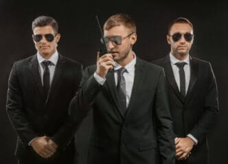 Three serious men in black suits and sunglasses, one speaking into a walkie-talkie, portraying a professional security team.