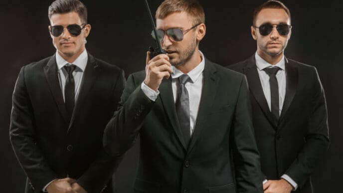 Three serious men in black suits and sunglasses, one speaking into a walkie-talkie, portraying a professional security team.
