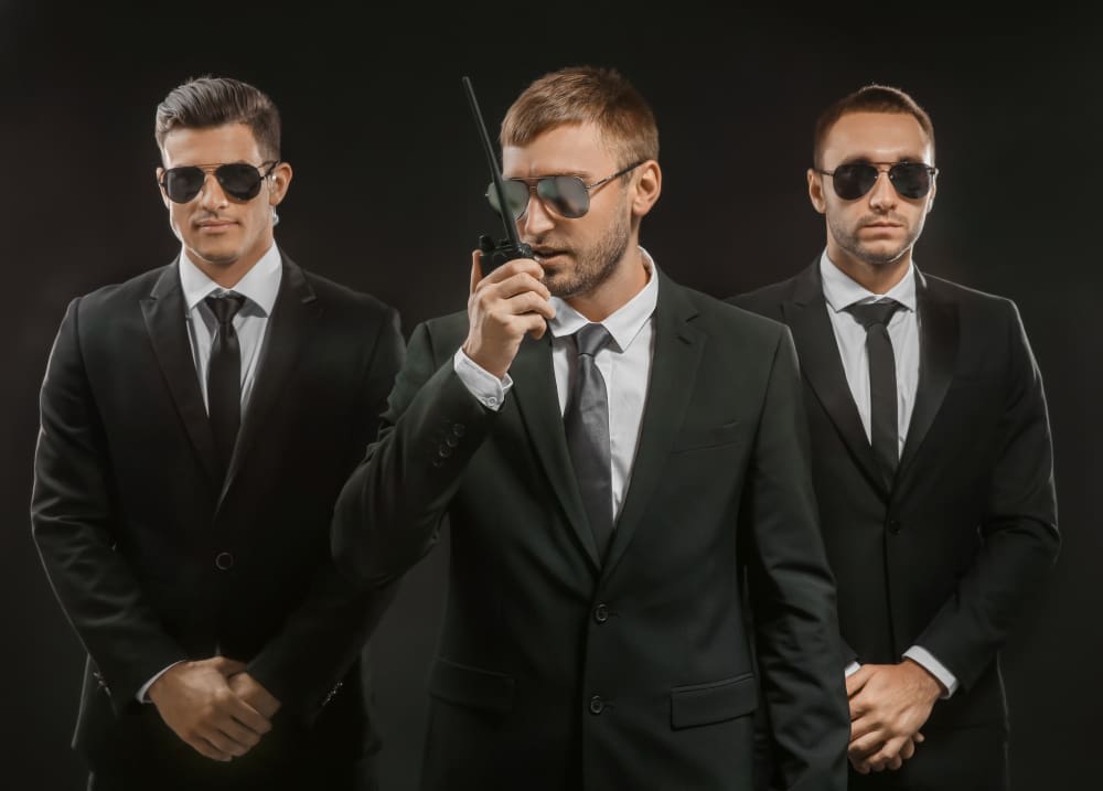 Three serious men in black suits and sunglasses, one speaking into a walkie-talkie, portraying a professional security team.