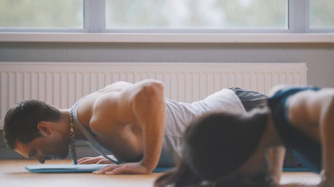 A man and a blurred woman performing push-ups on yoga mats in an indoor setting.