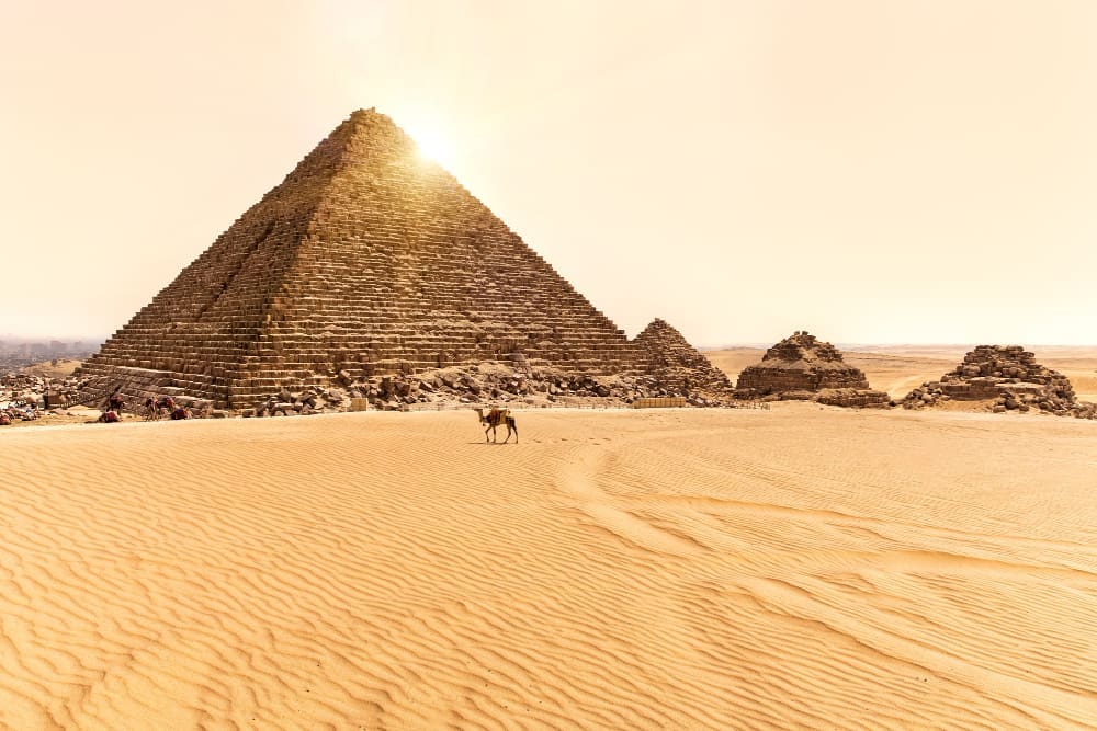 The Pyramid of Menkaure on the Giza Plateau, with smaller pyramids and a lone camel in the desert, under a bright sky.