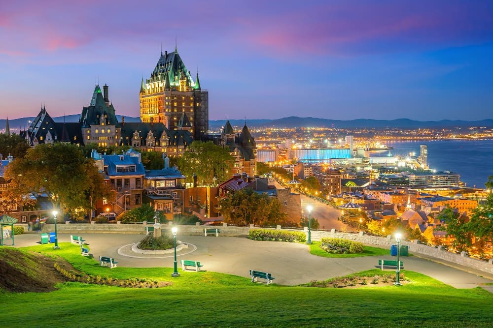 A panoramic view of Quebec City skyline at dusk, featuring the illuminated Château Frontenac hotel, other historic buildings, a park with glowing lampposts in the foreground, and the Saint Lawrence River with lights in the background.