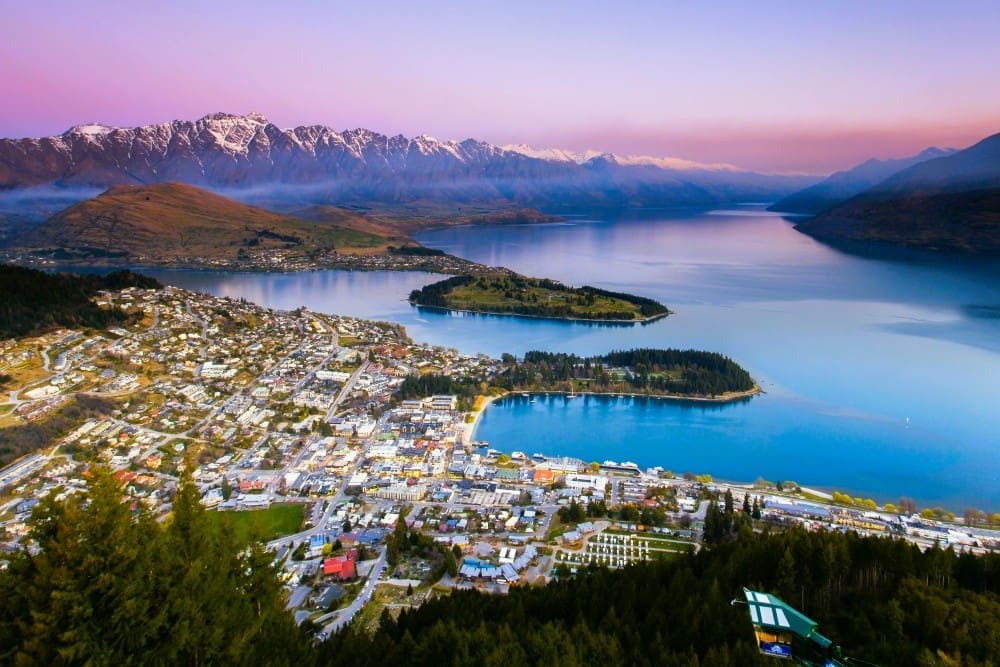 Aerial view of Queenstown, New Zealand, showcasing the town, Lake Wakatipu, and snow-capped Remarkables mountain range at twilight.