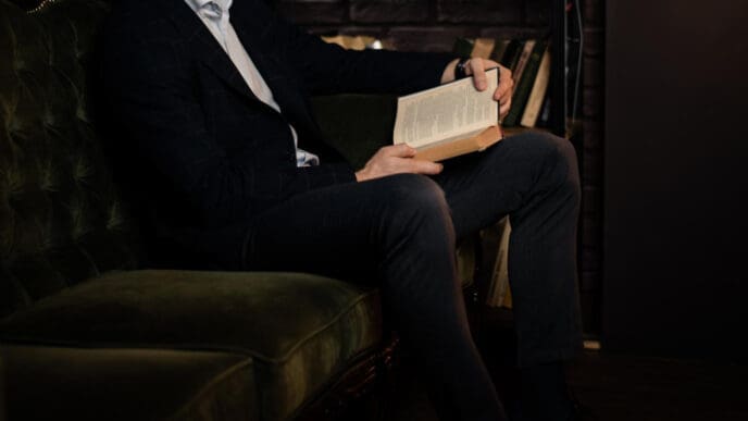 A man in a suit sitting on a velvet couch, holding an open book, with a bookshelf in the background, suggesting a moment of intellectual pursuit.