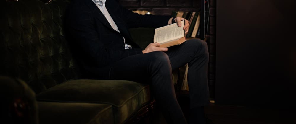 A man in a suit sitting on a velvet couch, holding an open book, with a bookshelf in the background, suggesting a moment of intellectual pursuit.