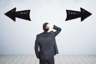 A man in a dark suit stands with his back to the viewer, scratching his head, facing a white wall with two large, hand-drawn black arrows pointing in opposite directions, symbolizing confusion or conflicting choices.