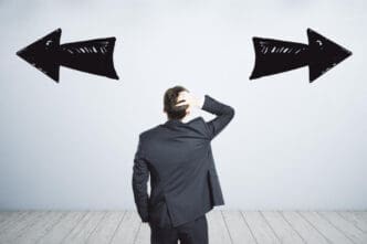 A man in a dark suit stands with his back to the viewer, scratching his head, facing a white wall with two large, hand-drawn black arrows pointing in opposite directions, symbolizing confusion or conflicting choices.