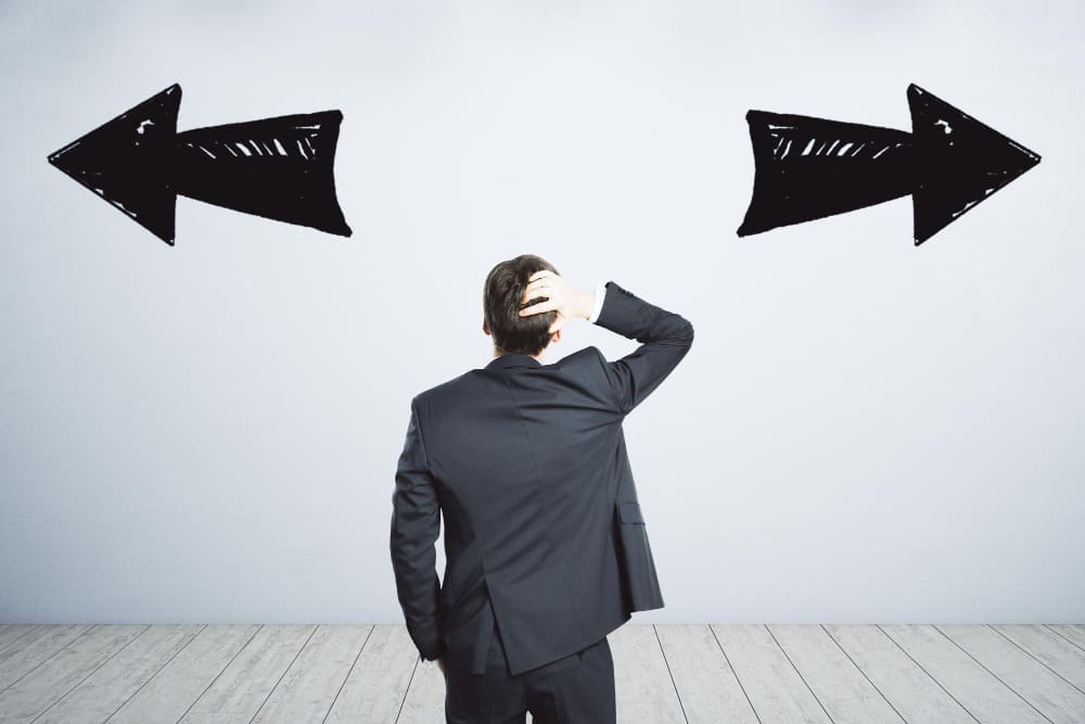 A man in a dark suit stands with his back to the viewer, scratching his head, facing a white wall with two large, hand-drawn black arrows pointing in opposite directions, symbolizing confusion or conflicting choices.
