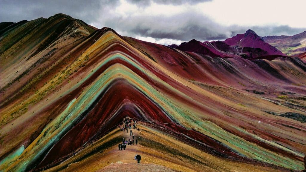 A panoramic view of Peru's Rainbow Mountain (Vinicunca), showing its vibrant stripes of colored earth and hikers on its path under a cloudy sky.