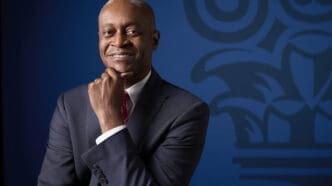 A professional portrait of Raphael Bostic, smiling warmly and posing with his hand under his chin, wearing a dark suit and red tie against a blue background with a blurred institutional logo.