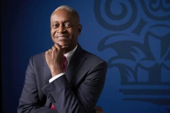 A professional portrait of Raphael Bostic, smiling warmly and posing with his hand under his chin, wearing a dark suit and red tie against a blue background with a blurred institutional logo.
