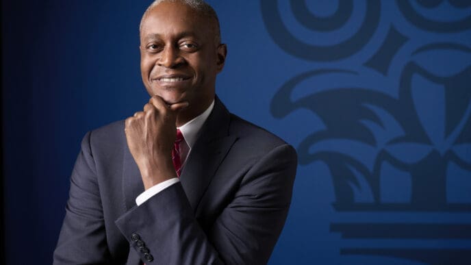 A professional portrait of Raphael Bostic, smiling warmly and posing with his hand under his chin, wearing a dark suit and red tie against a blue background with a blurred institutional logo.
