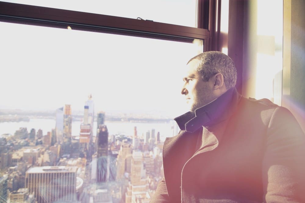 A man in a coat looking out a large window at a sprawling city skyline and bright sky.