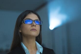 A focused woman wearing glasses reflecting blue light, looking upwards, with a projector beam in the background, symbolizing forward thinking and digital engagement.