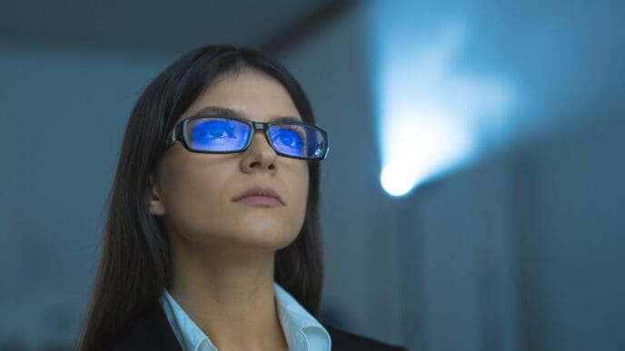 A focused woman wearing glasses reflecting blue light, looking upwards, with a projector beam in the background, symbolizing forward thinking and digital engagement.