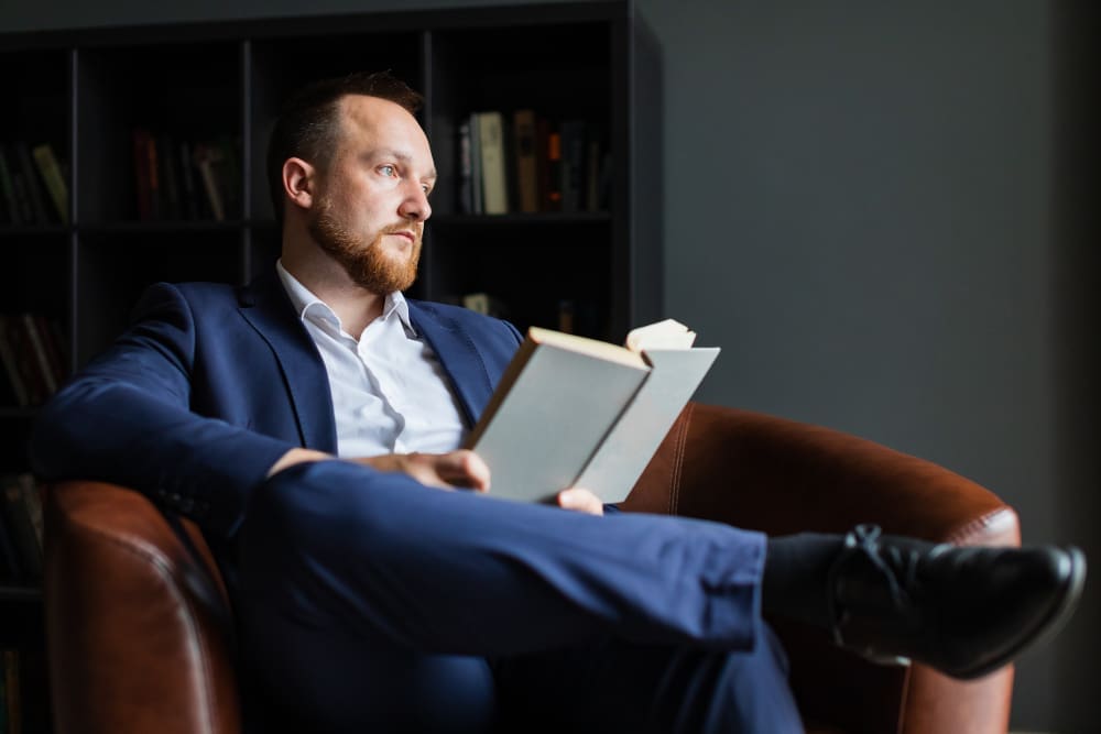 A successful businessman with a beard, wearing a blue suit, sitting in a leather armchair and looking thoughtfully away from the book he is holding.