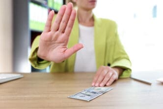 A person's hand raised in a stop gesture towards a hundred-dollar bill on a desk.