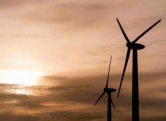 Silhouettes of two wind turbines stand against a warm, cloudy sunset sky, with the sun low on the horizon.